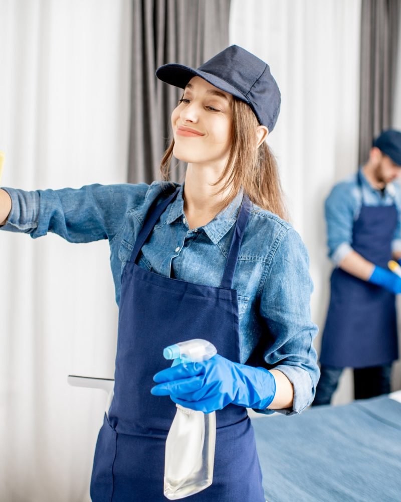 Professional cleaners during the work indoors Couple as a professional cleaners in uniform rubbing furniture and wiping dust in the bedroom or hotel room. Cleaning service concept